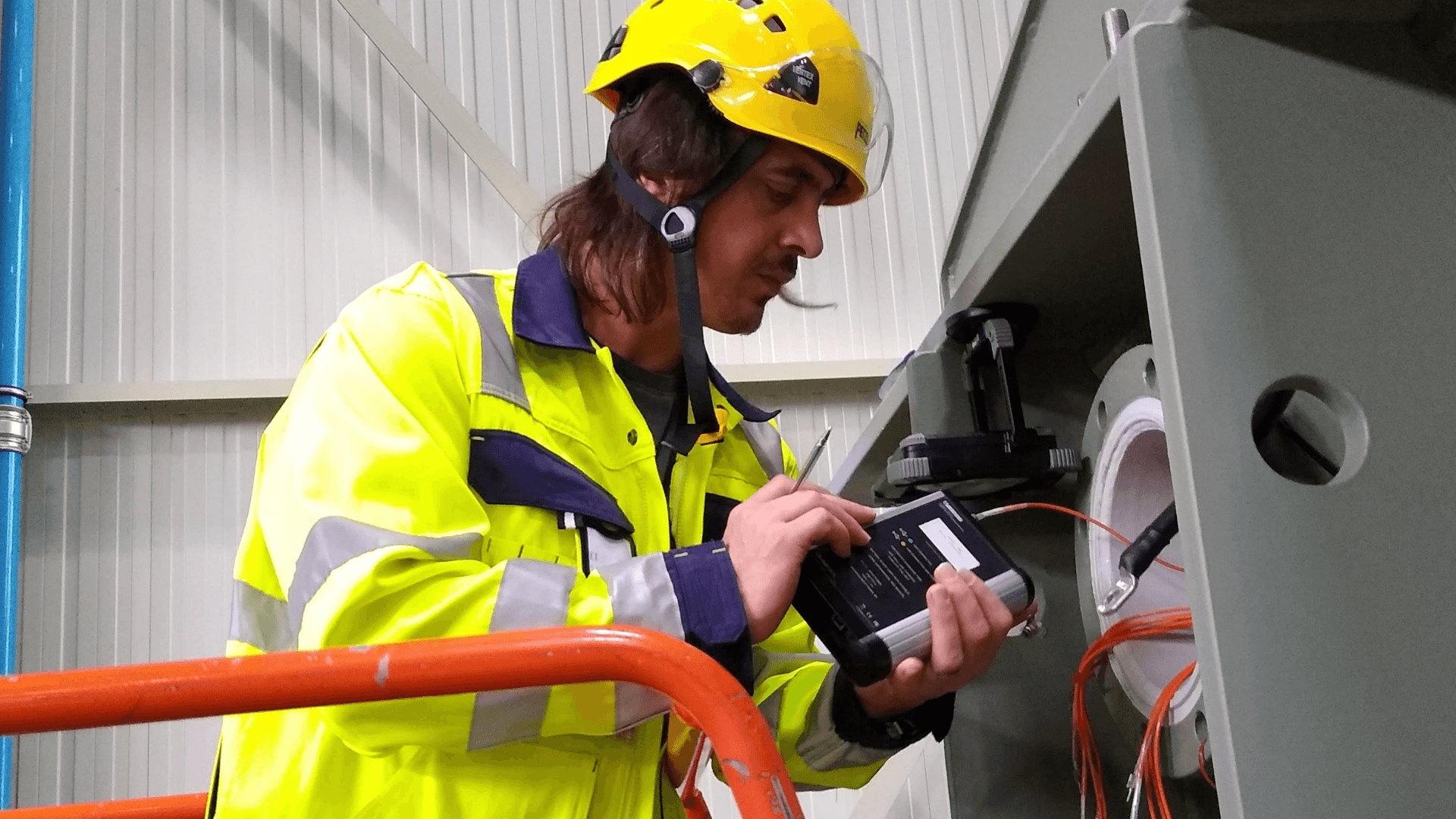 Technician in yellow hard hat and reflective vest testing electrical equipment with diagnostic device